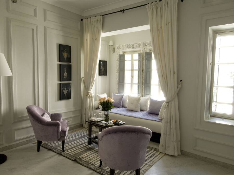 Una habitación con una cama, dos sillones, una lámpara y un ventanal con cortinas.