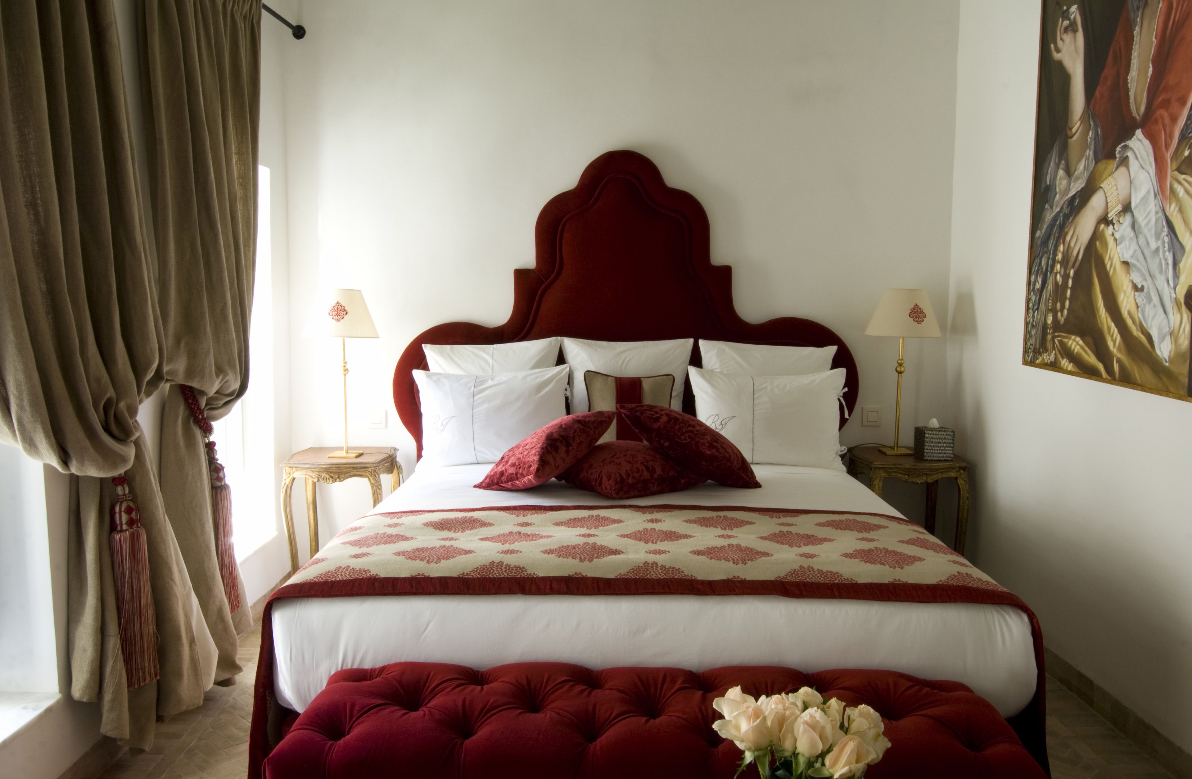 Un lit bien fait, avec une tête de lit rouge, une literie blanche et un couvre-lit à motifs rouges et blancs. On y trouve deux tables de chevet avec des lampes, une grande fenêtre avec des rideaux et un tableau accroché au mur.
