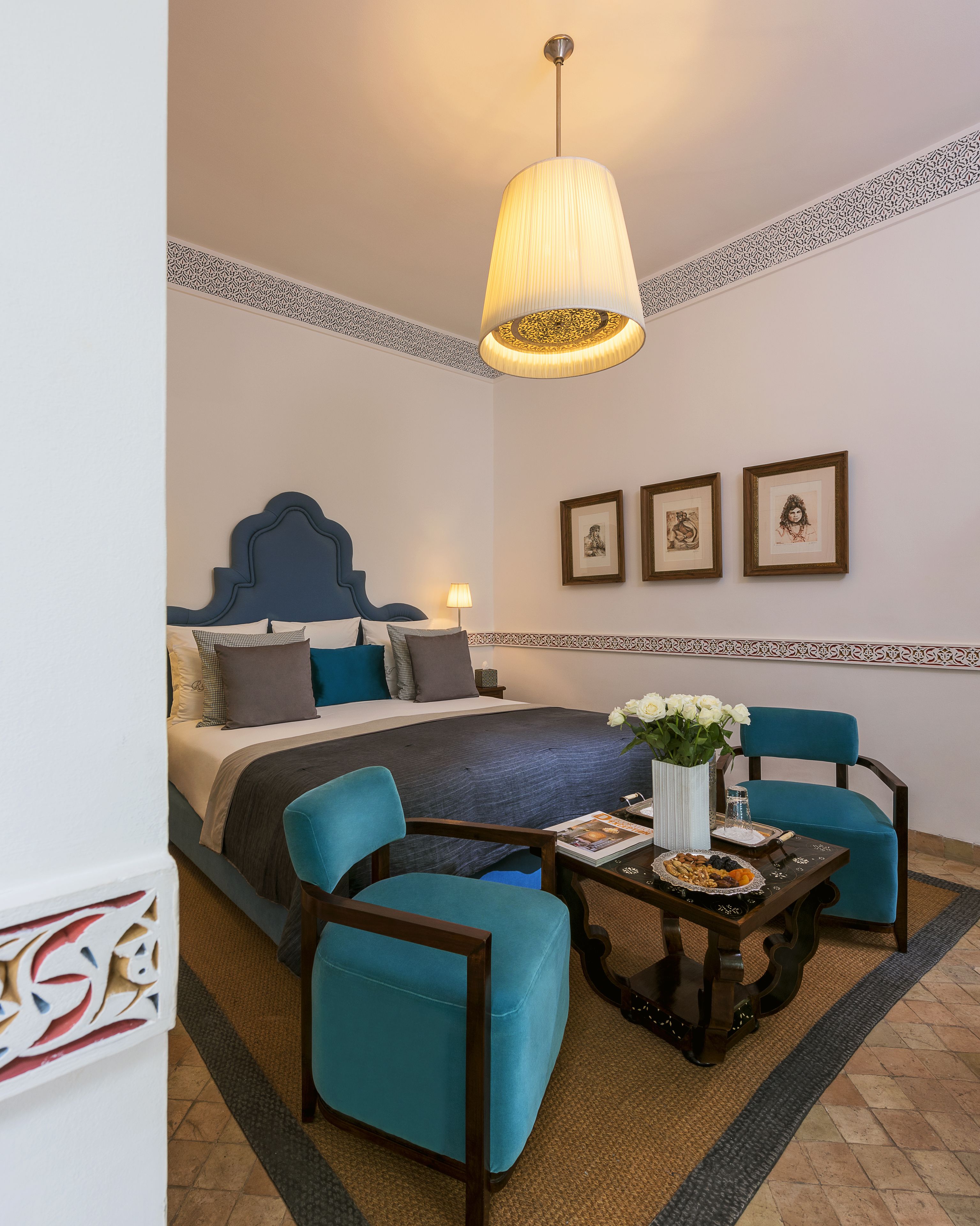 Un cuarto de hotel con una cama, dos sillones azules, una mesa de centro con un florero y tres cuadros en la pared.