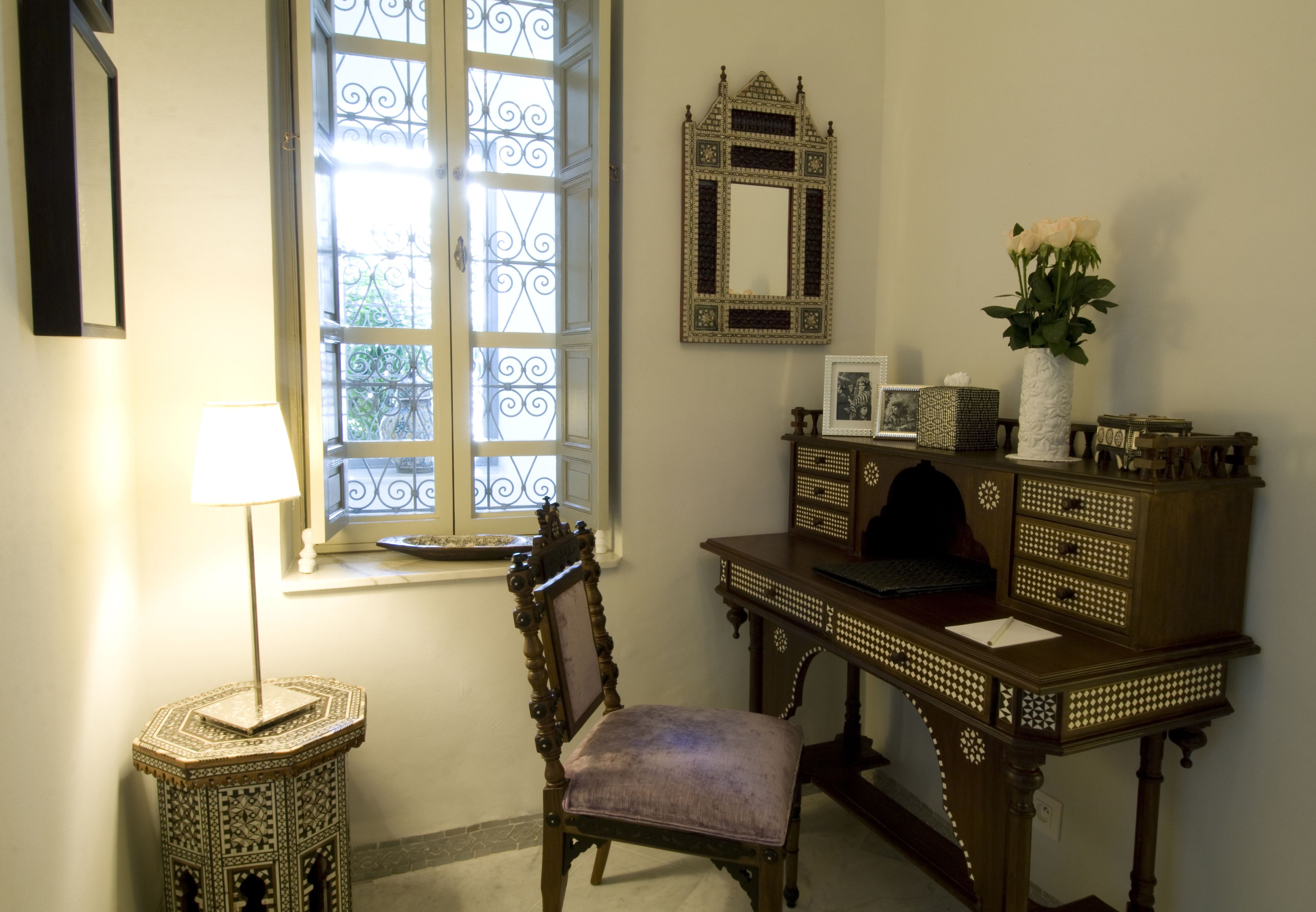 A room with a traditional wooden desk, a chair, a decorative mirror, and a window with iron bars.