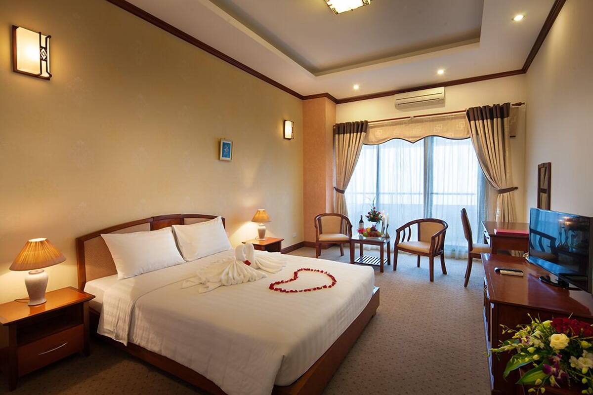 Quarto de hotel com uma cama grande, uma mesa com televisão, uma cadeira, uma mesinha com flores e uma janela com cortinas.