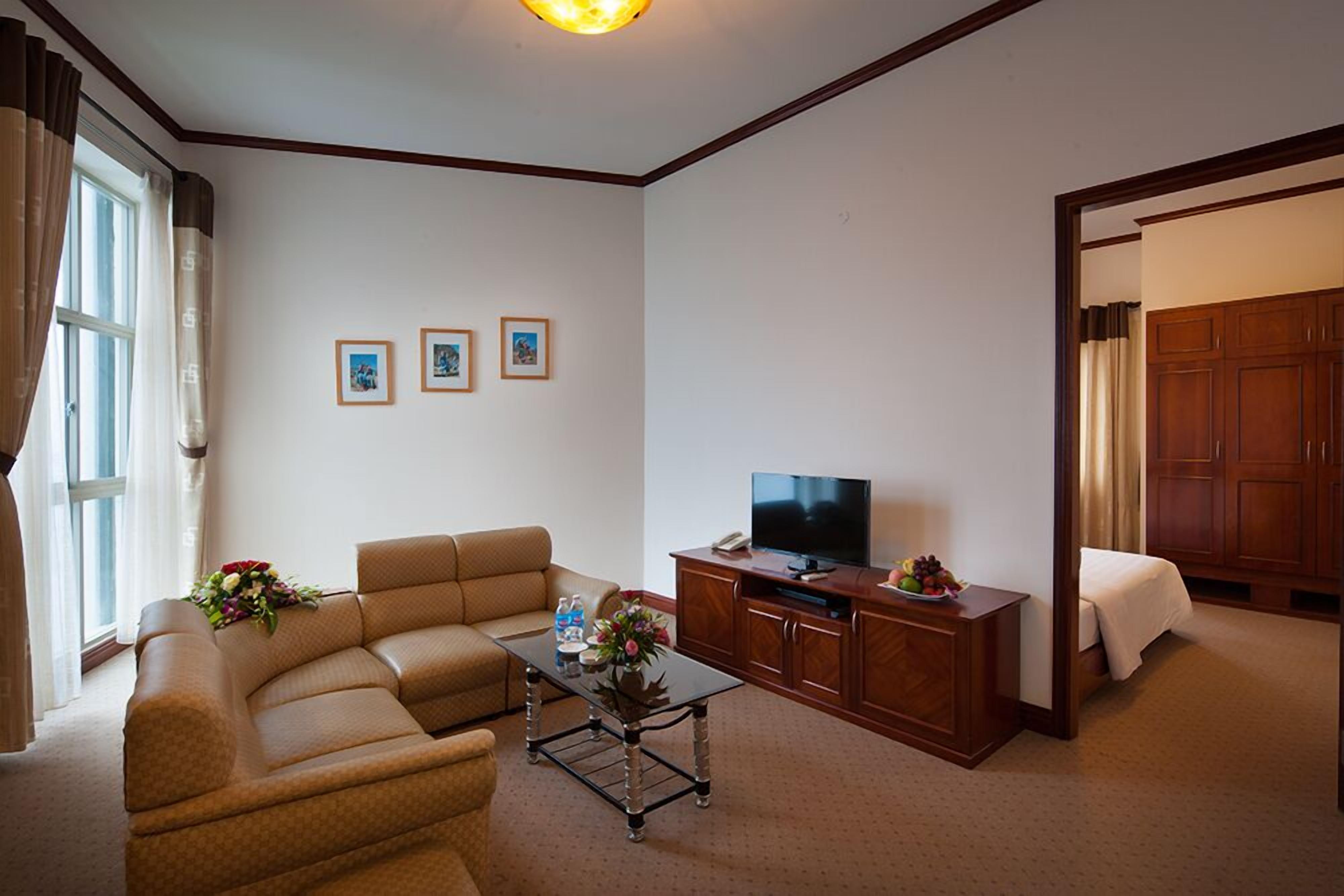 Une chambre d’hôtel avec un canapé, une télévision, une petite table avec un vase et des fleurs, et un lit doté d’une tête de lit en bois.