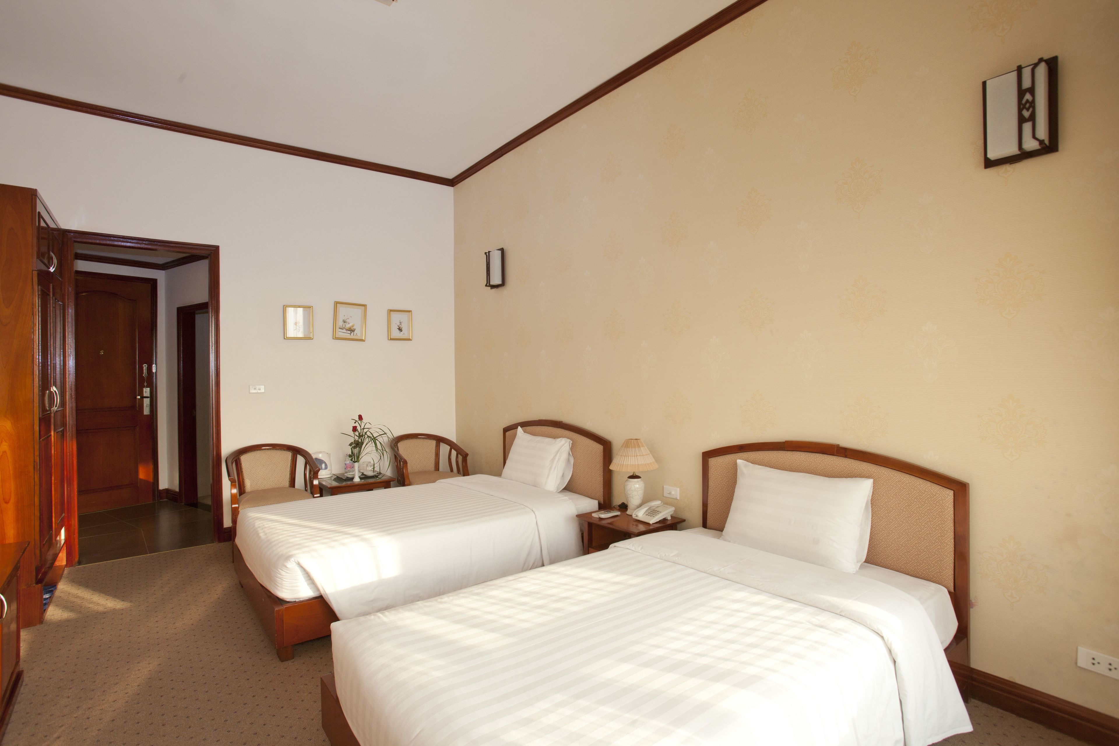 Quarto de hotel com duas camas, um guarda-roupa de madeira, uma escrivaninha com luminária e uma mesinha de centro com um vaso.