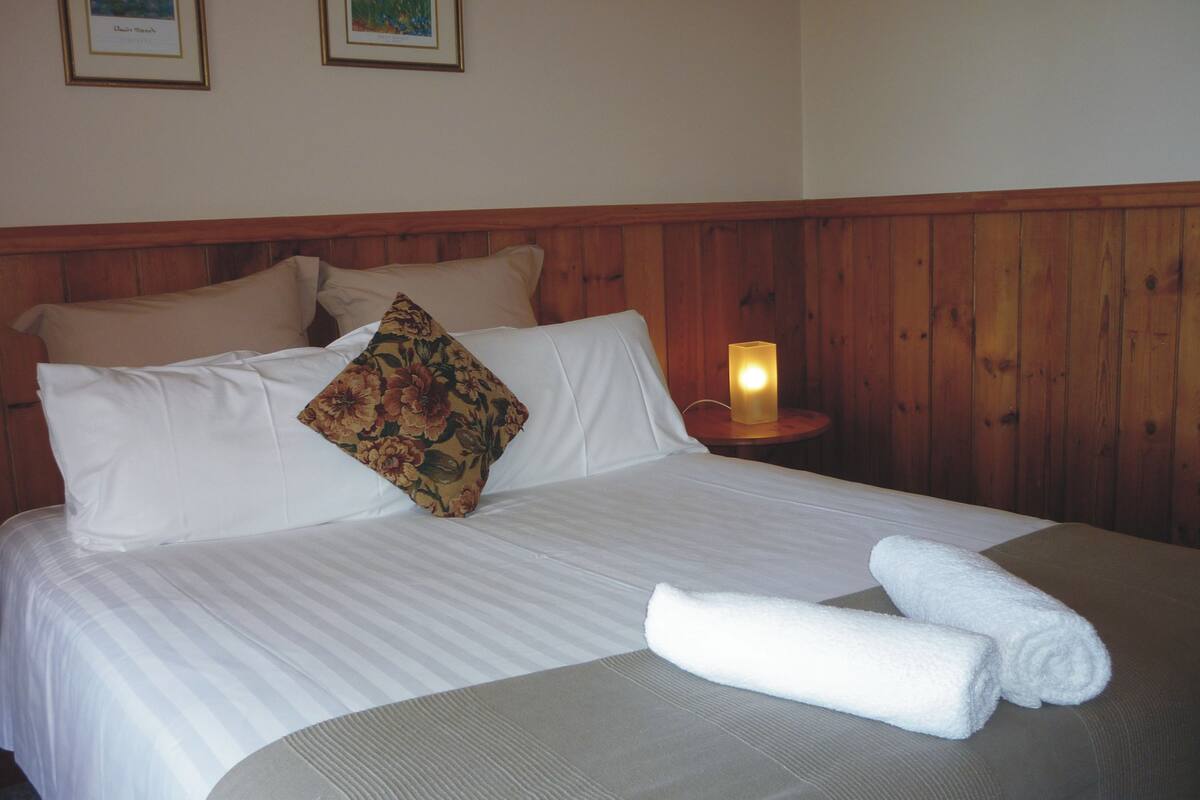 Cama bem arrumada com lençóis brancos, um travesseiro floral e uma toalha enrolada em uma cabeceira de madeira.