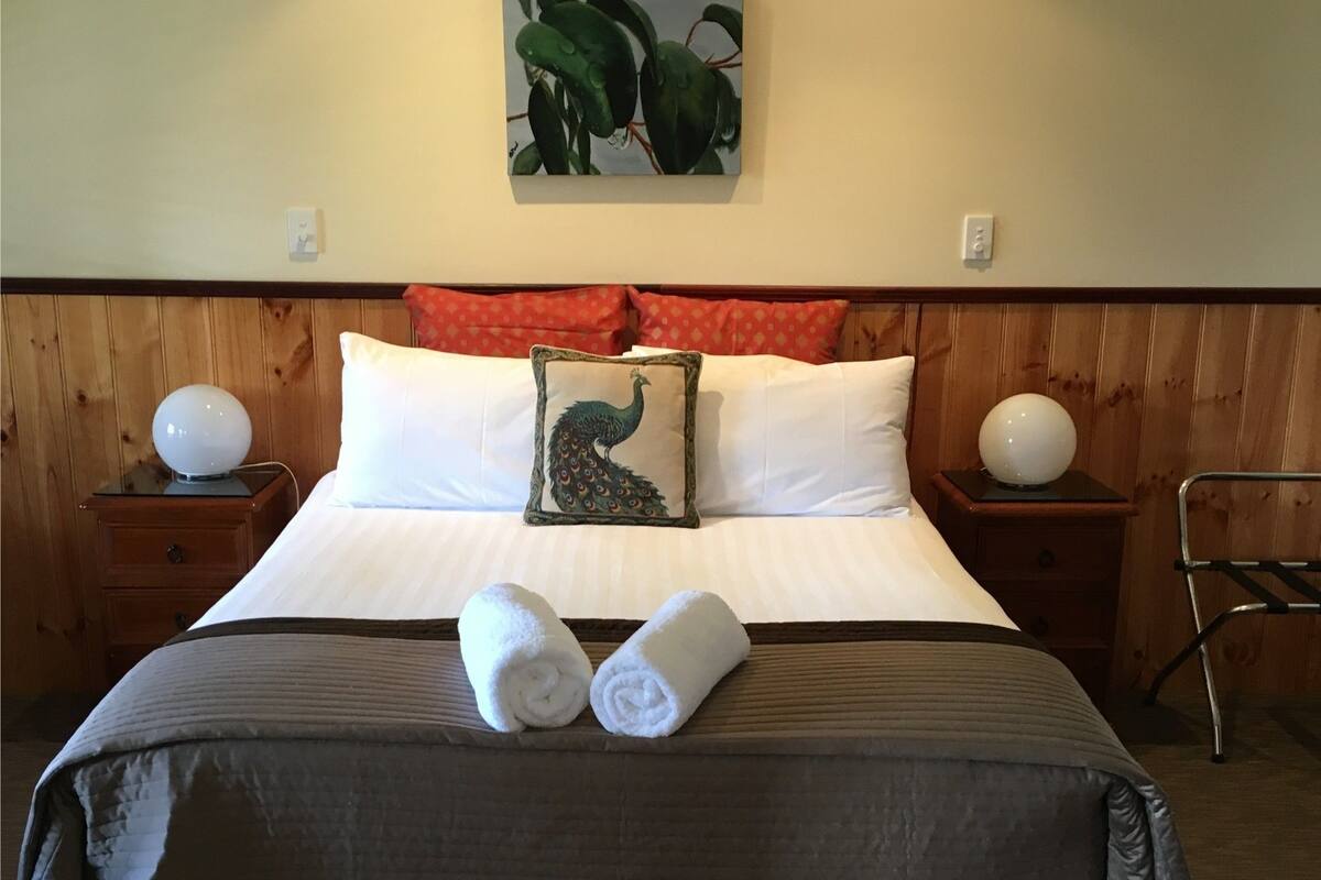 Uma cama bem arrumada com lençóis brancos e um travesseiro decorativo, duas mesas de cabeceira com luminárias, um quadro pendurado na parede e uma cabeceira de madeira.