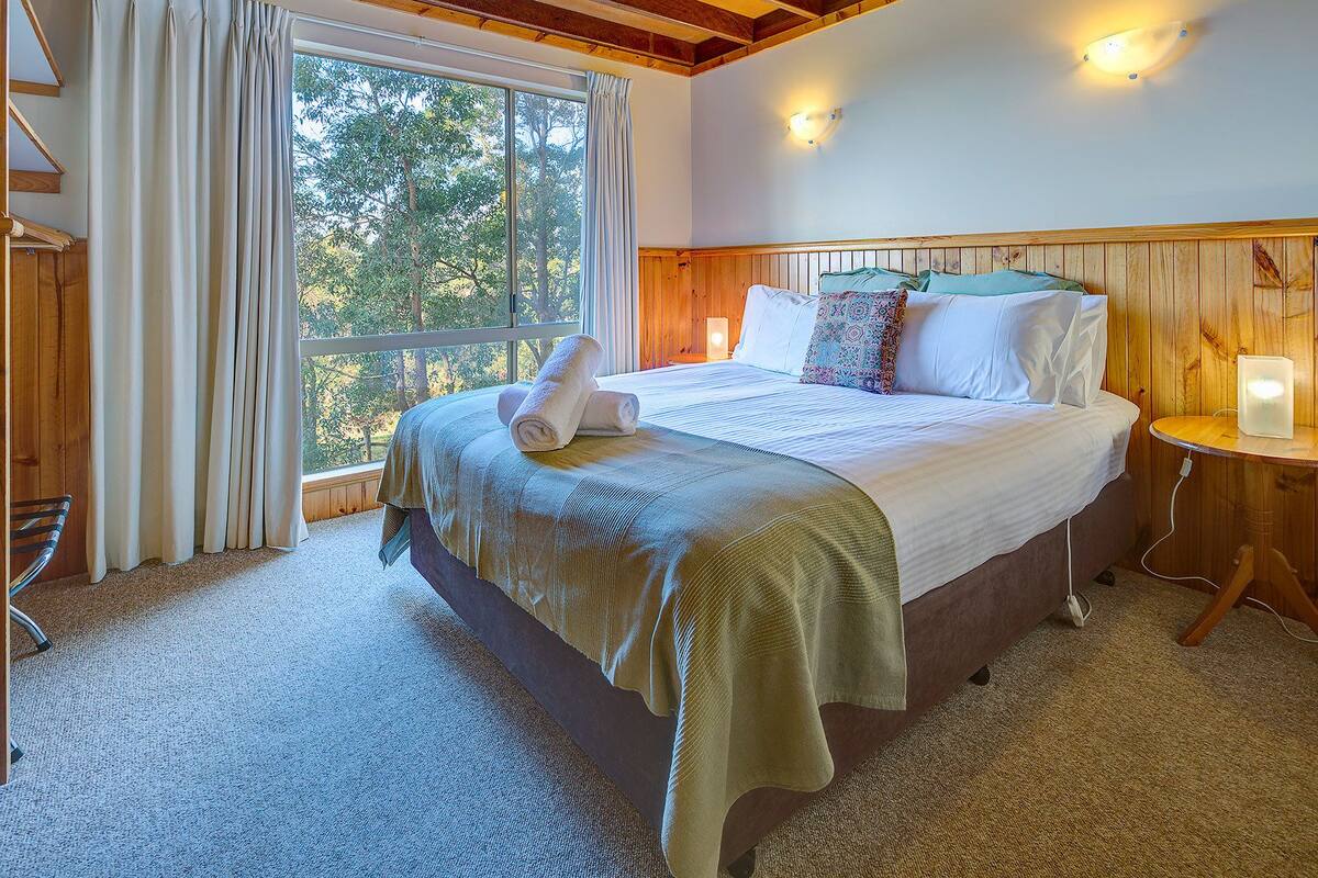 Quarto com cama grande, mesa de cabeceira, janela com cortinas e vista para árvores.