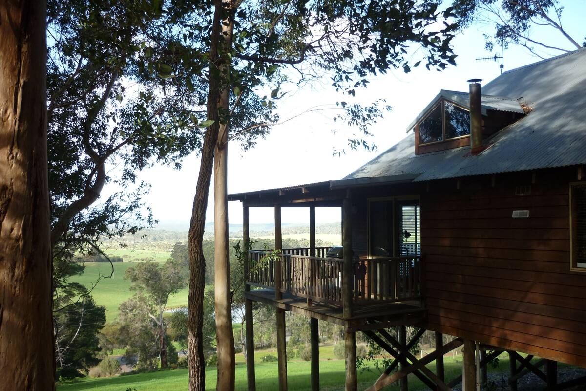 Uma cabana de madeira com varanda, rodeada por árvores e vista para uma paisagem verde.