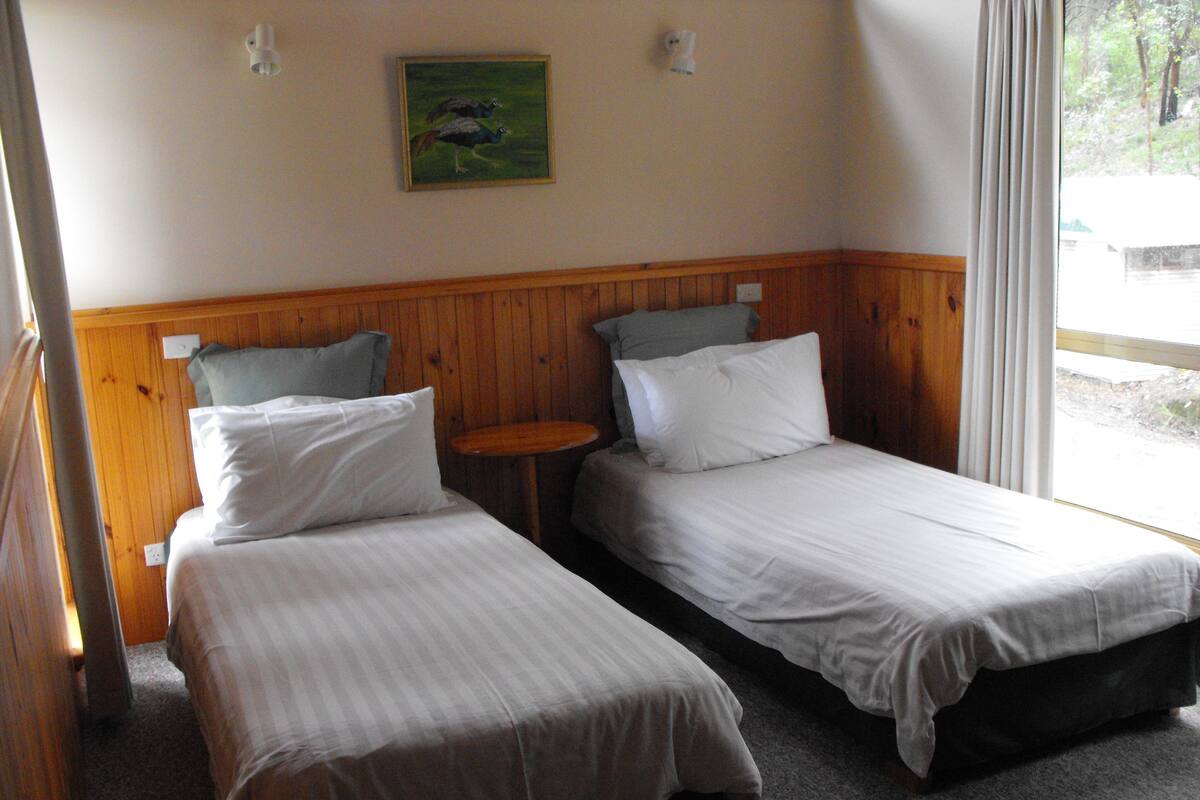 Quarto com duas camas de solteiro, painéis de madeira nas paredes, uma mesa redonda pequena e um quadro de pássaros na parede.