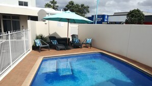 Piscina con borde azulejado, rodeada por una cerca blanca. Se observan sillas de descanso con cojines azules y una sombrilla azul.
