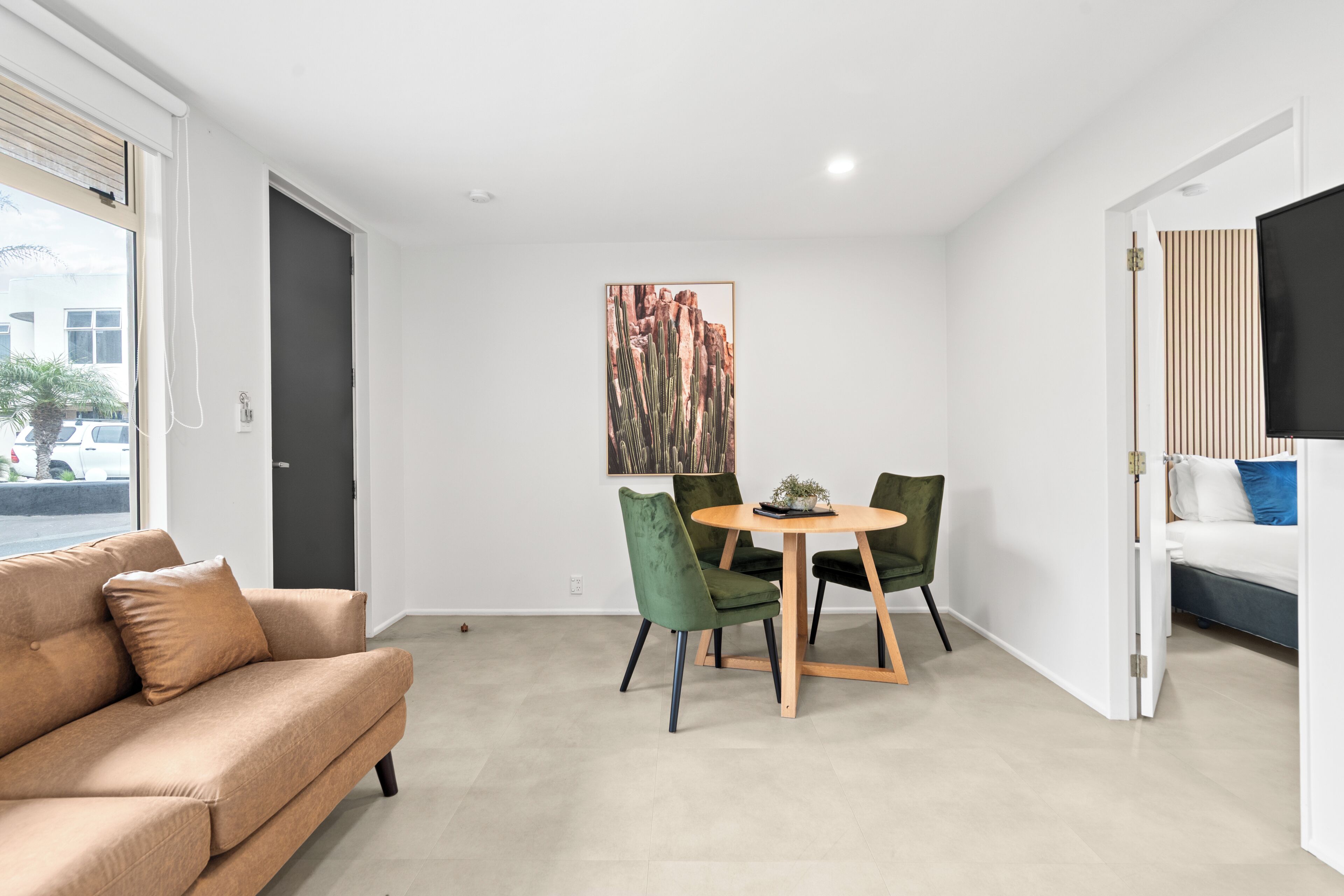A modern living room with a beige sofa, a dining table with chairs, a painting on the wall, and a bed with a TV.