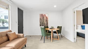 A modern living room with a beige sofa, a dining table with chairs, a painting on the wall, and a bed with a TV.