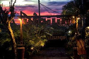 Une enseigne portant le nom « NELUWUNGAAN », dans un décor tropical, avec un coucher de soleil en arrière-plan.