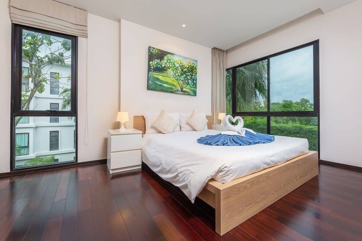 Une chambre moderne avec un grand lit, une table de chevet, un tableau et une vue sur la verdure grâce à de grandes fenêtres.