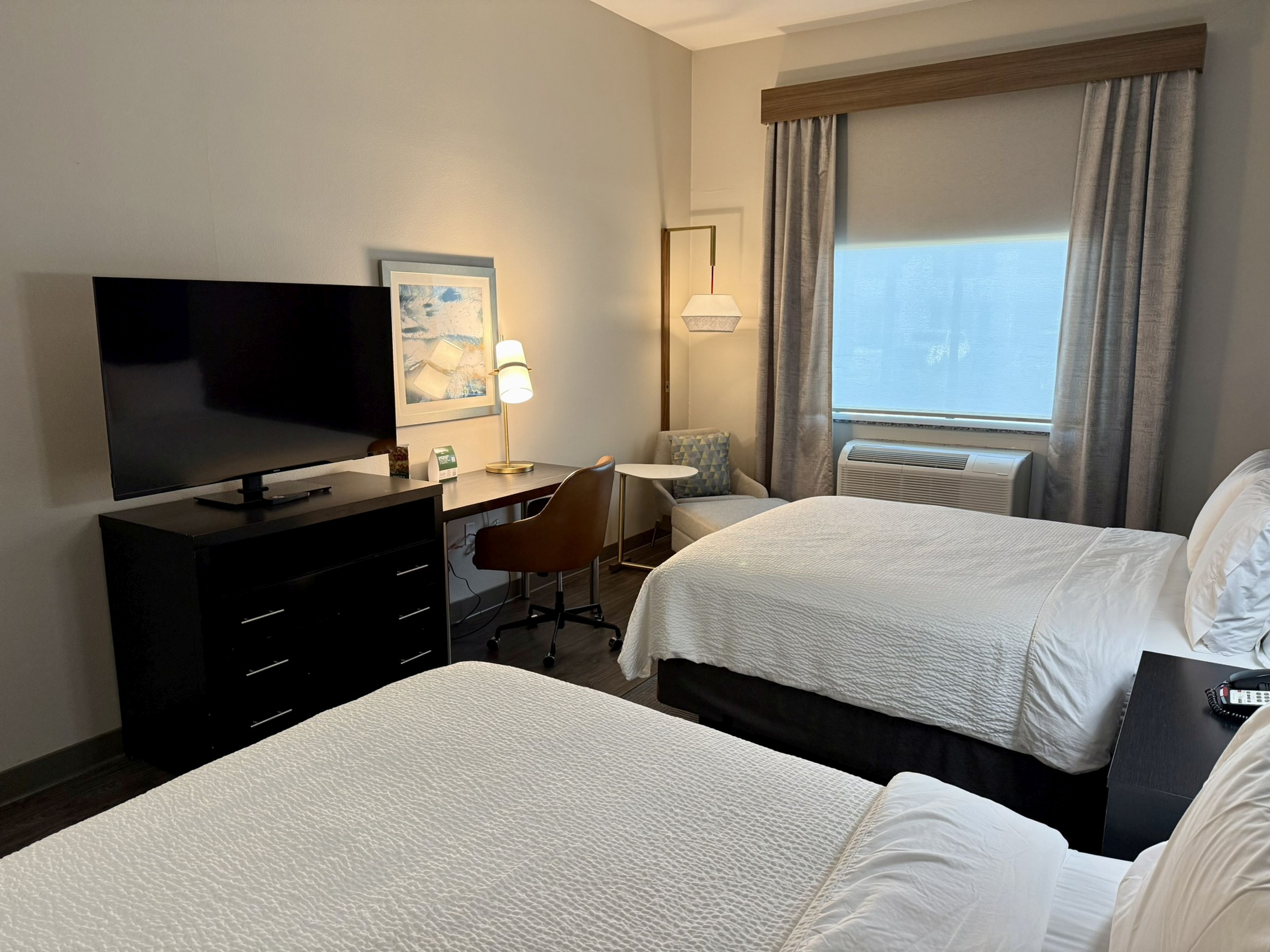 Habitación de hotel con dos camas, un escritorio con silla, televisor y ventana con cortinas.
