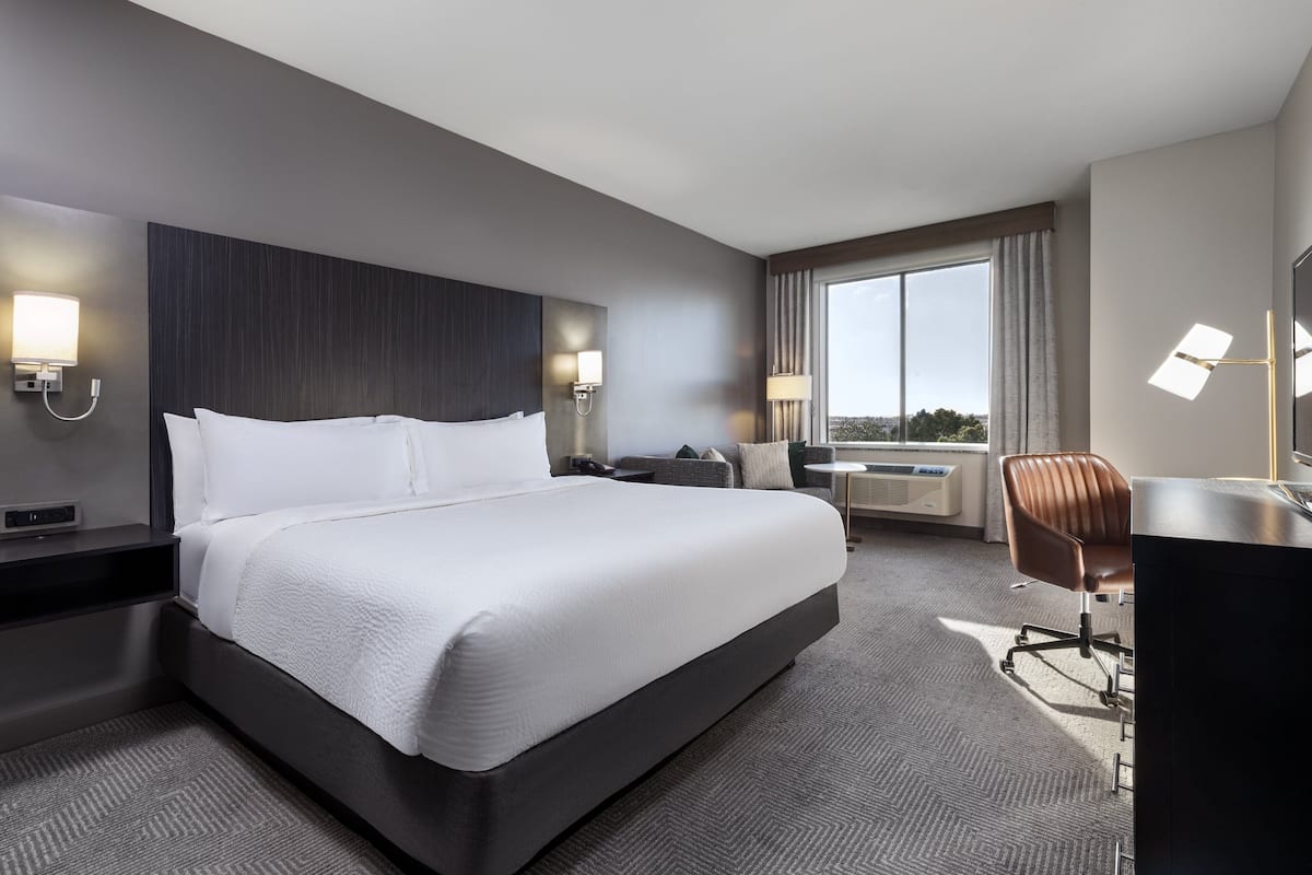 A modern hotel room with a large bed, a desk, a chair, and a view of the outdoors through a large window.