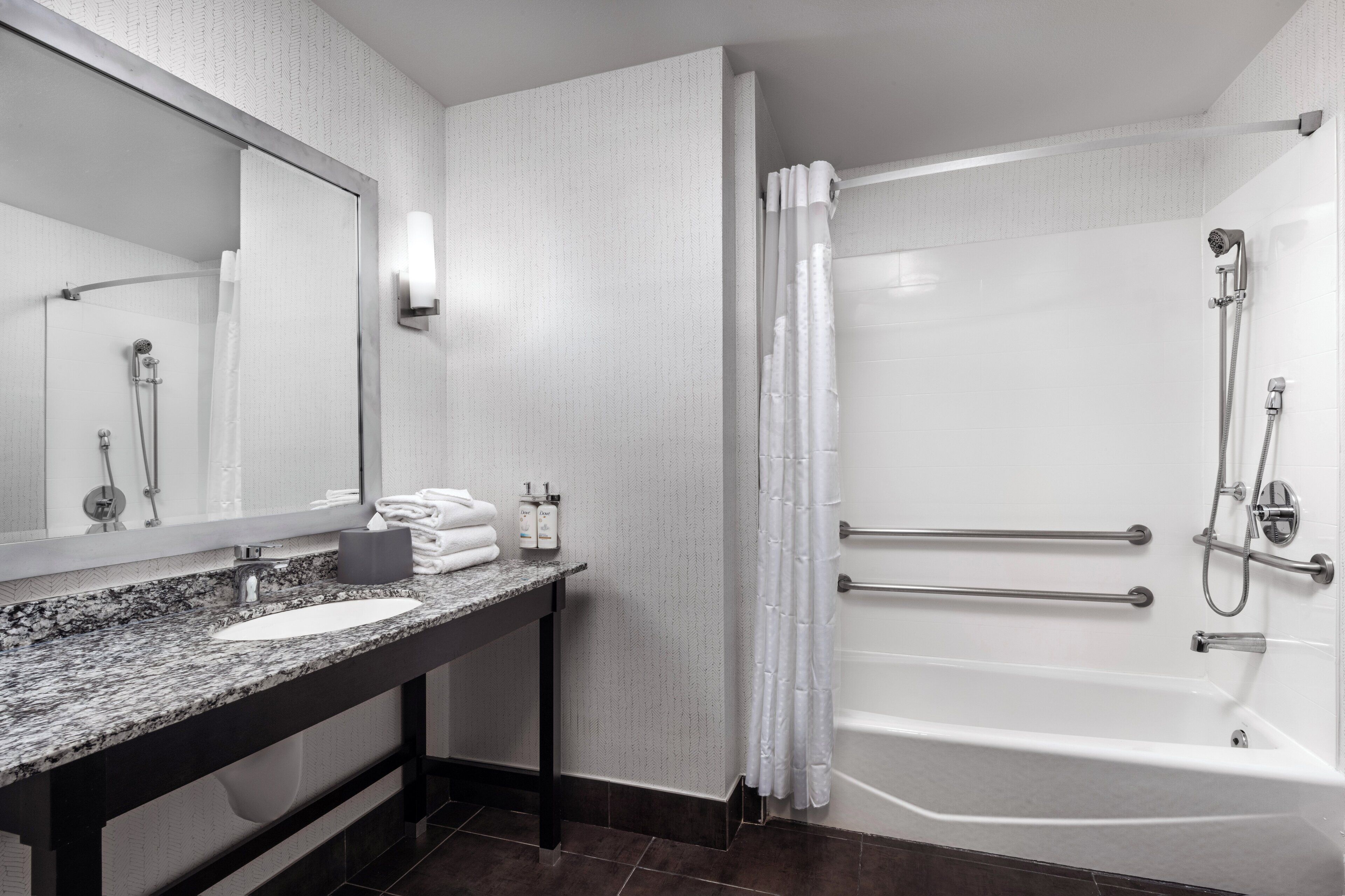 A modern bathroom with a granite countertop sink, a large mirror, a bathtub with grab bars, and a shower with a handheld showerhead.