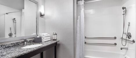 A modern bathroom with a granite countertop sink, a large mirror, a bathtub with grab bars, and a shower with a handheld showerhead.