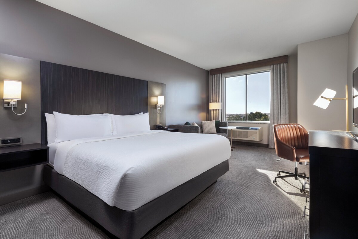 Een moderne hotelkamer met een groot bed, een bureau, een stoel en uitzicht op het landschap door een groot raam.