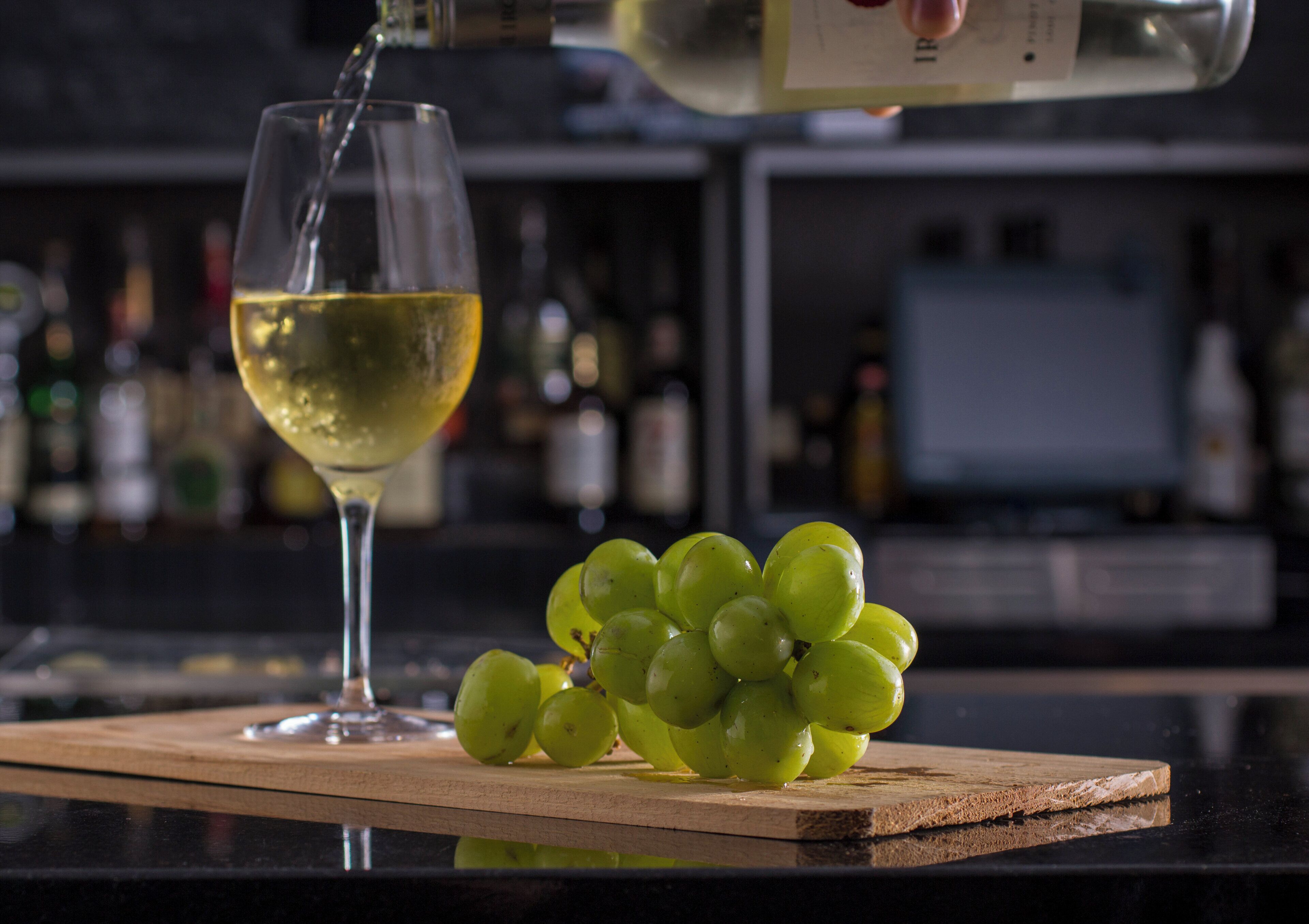 Ein Glas Weißwein auf einer Holzplatte mit einer Traube grüner Weintrauben daneben.