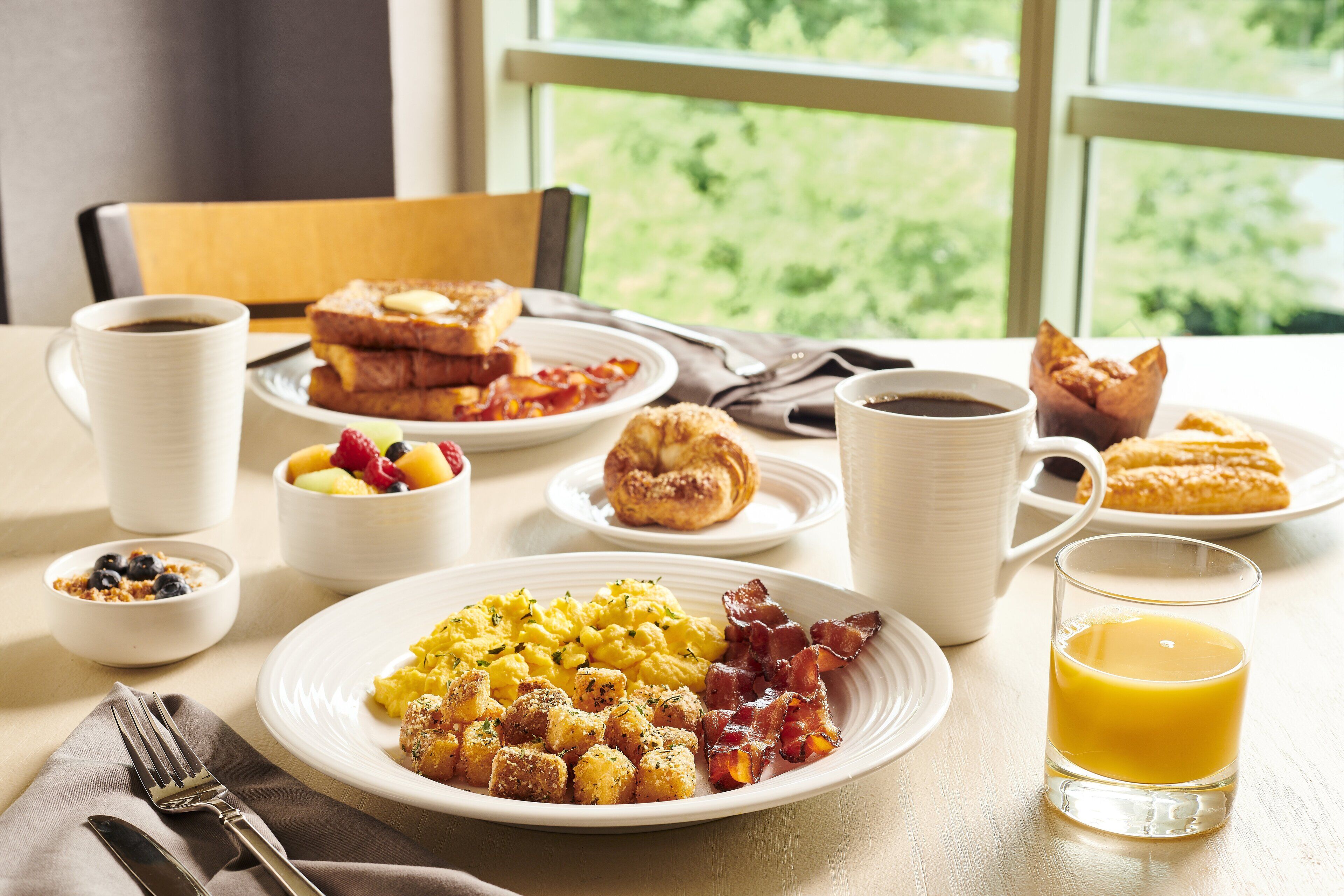 Una mesa de desayuno con platos de huevos revueltos, tocino, tostadas y pasteles, junto con tazas de café y jugo.