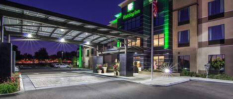 Un hotel Holiday Inn con entrada cubierta, estacionamiento exterior e iluminación frontal destacada.