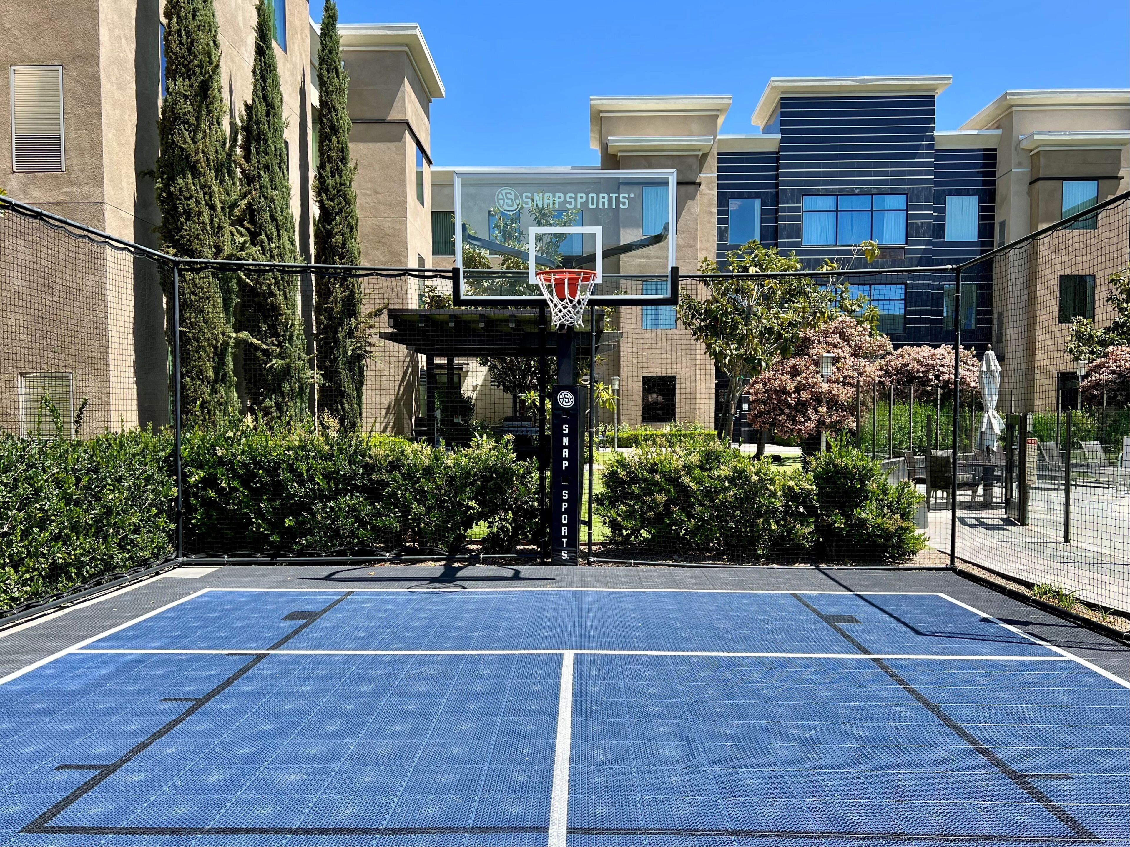 An outdoor basketball court with a net, surrounded by a fence and greenery.