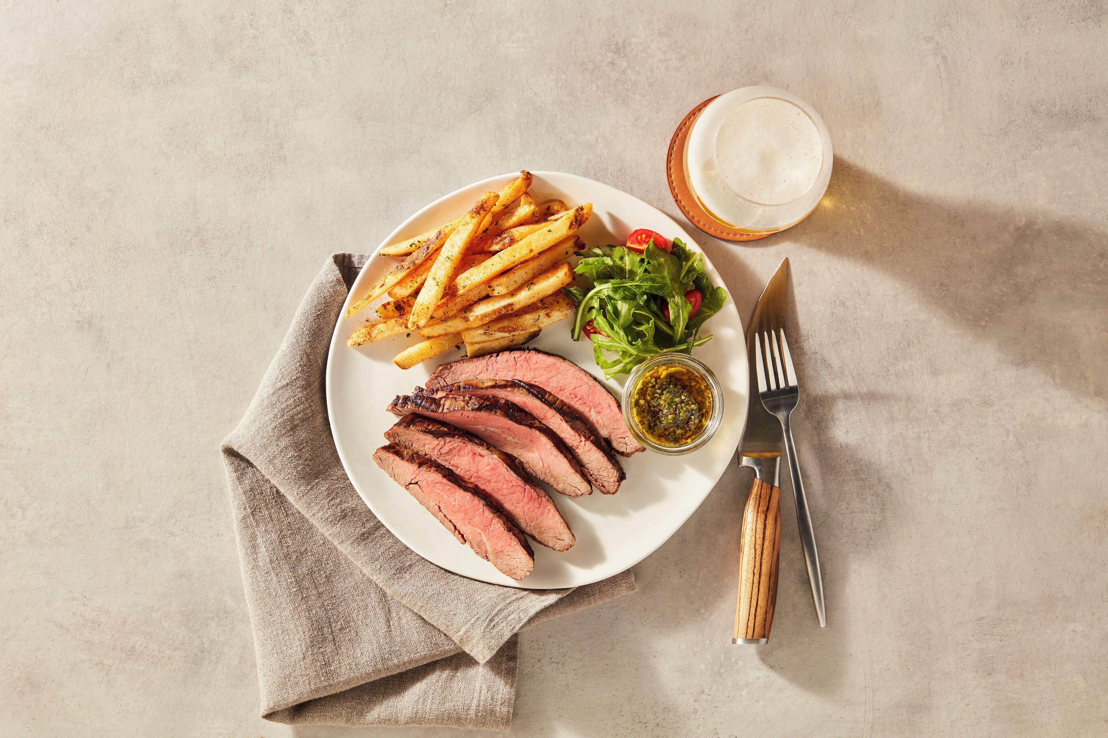 Un plato de comida que incluye bistec, papas fritas, ensalada y una bebida, sobre una mesa con servilleta y cubiertos.