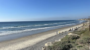 Uma praia com costa rochosa, céu claro e algumas pessoas caminhando pela trilha.
