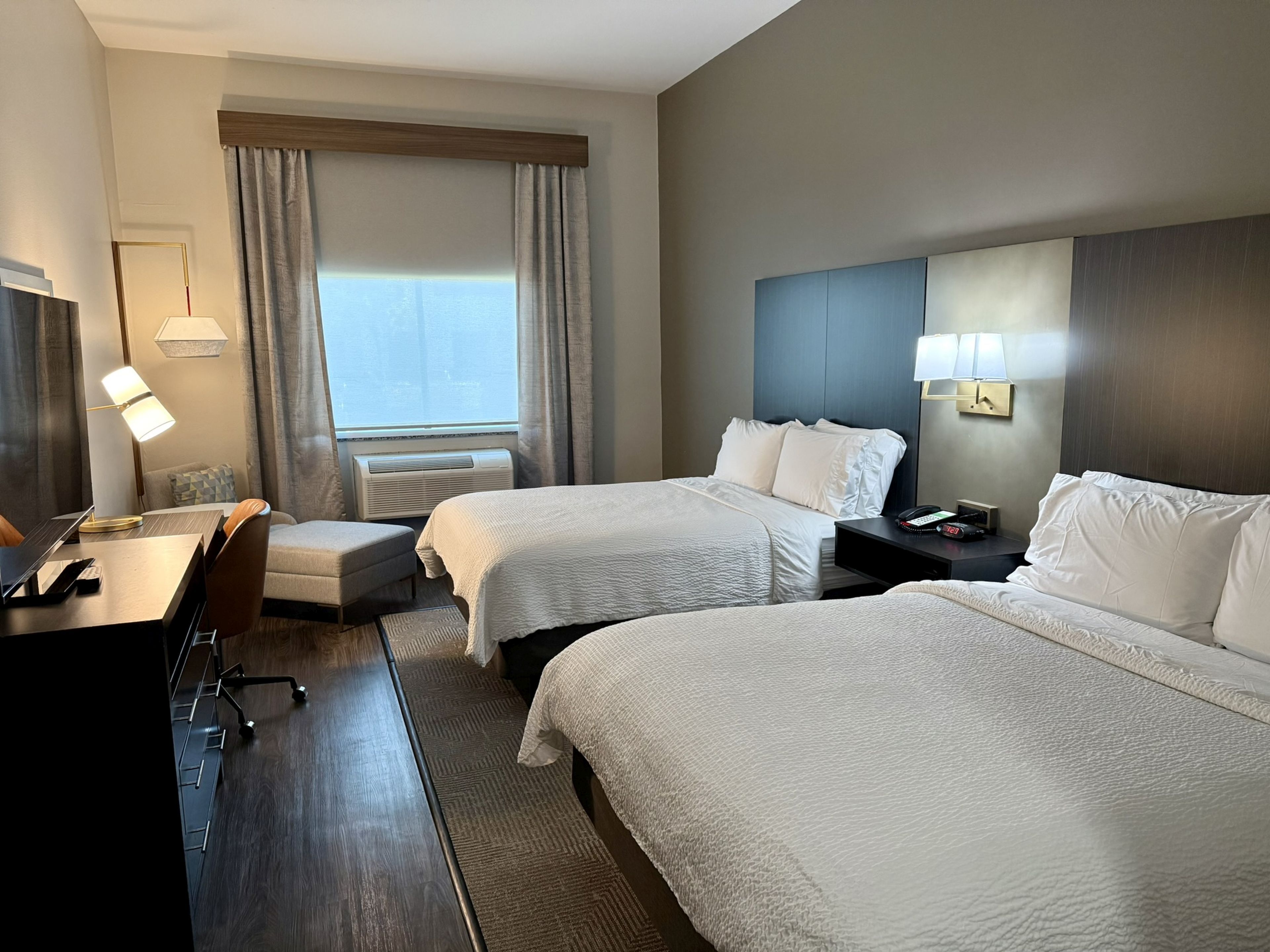 Habitación de hotel con dos camas, un escritorio, una silla, una lámpara y un ventanal con cortinas.