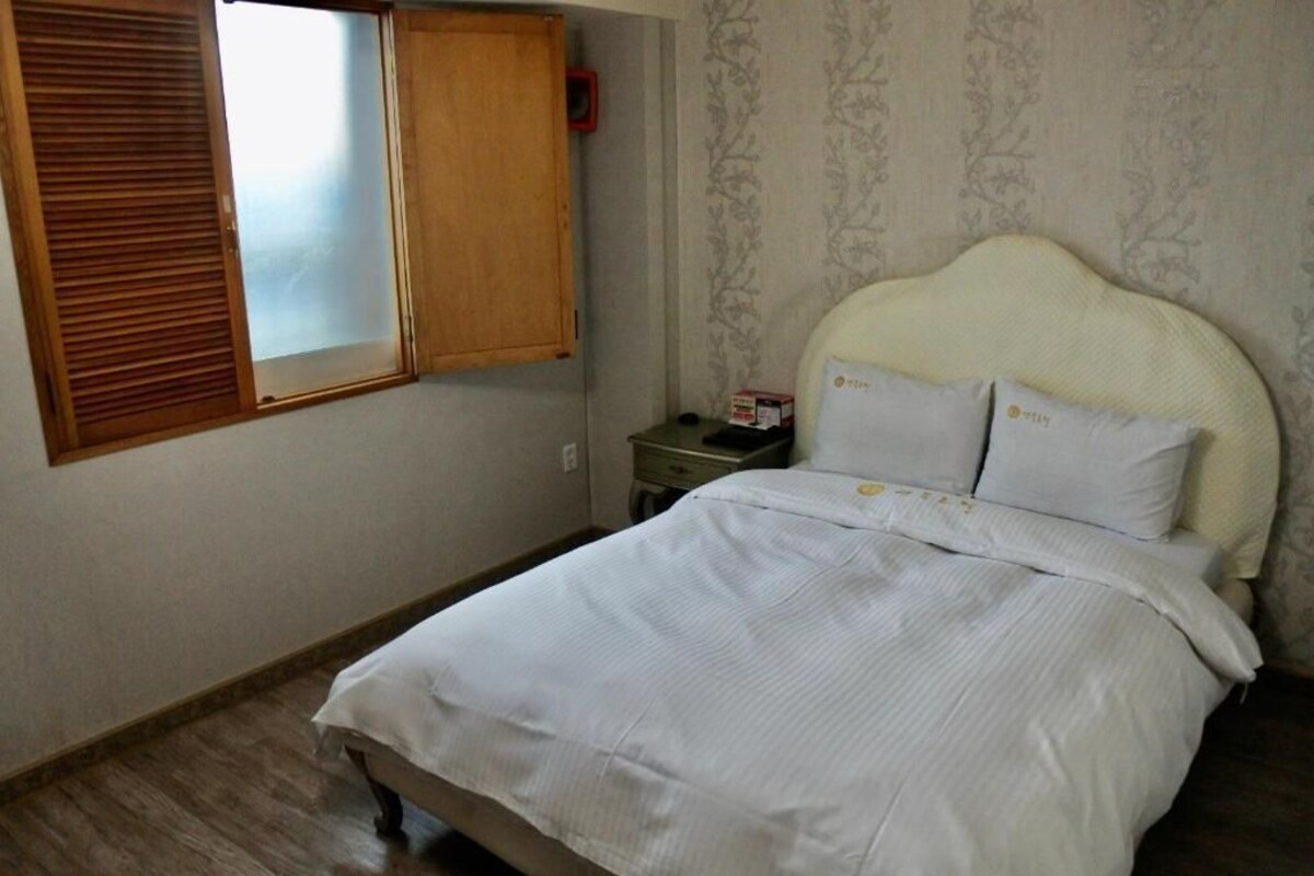 A bedroom with a bed, a nightstand, a wooden wardrobe, and a window with blinds.