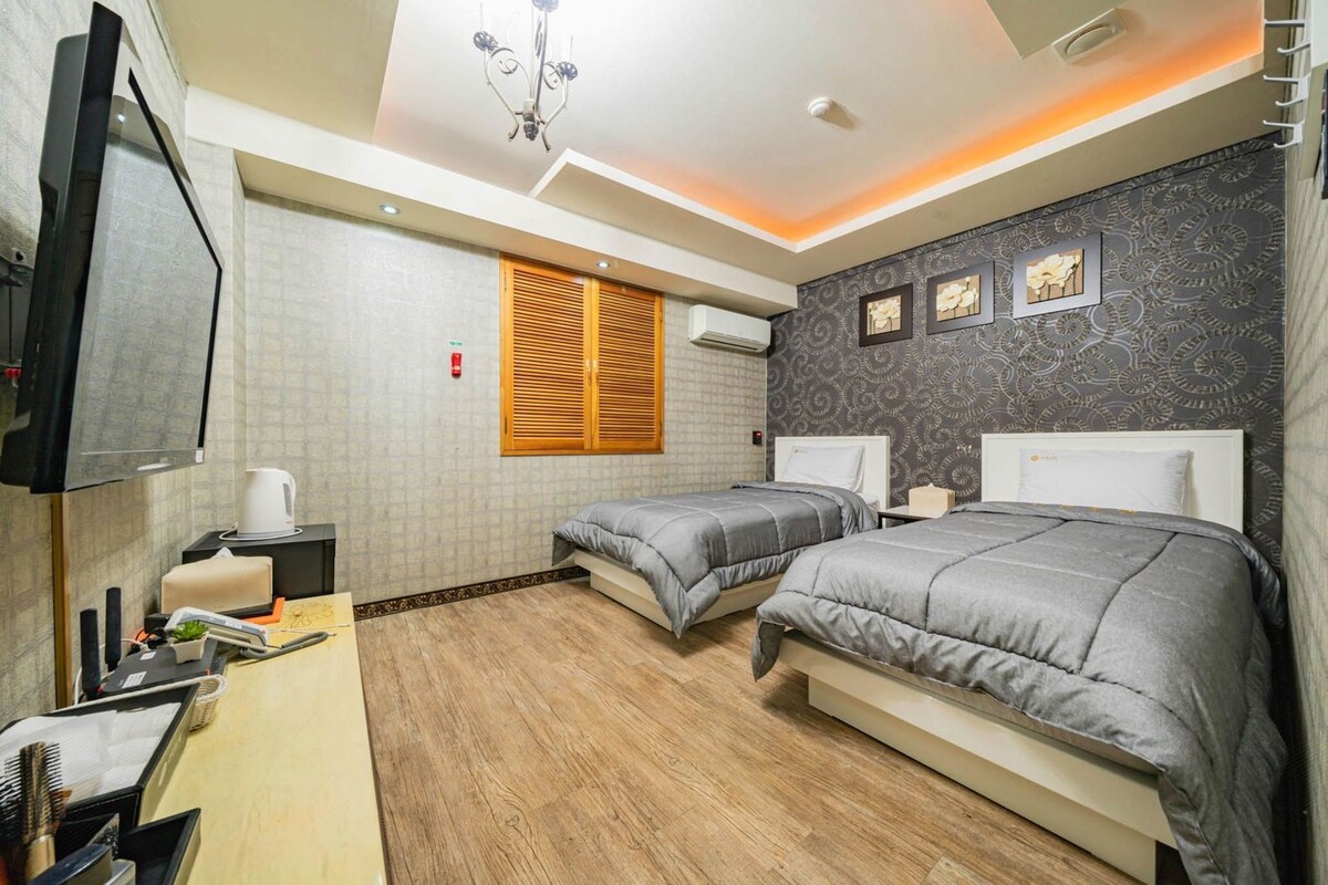 A hotel room with two beds, a desk with a kettle, a TV, and a patterned wall.