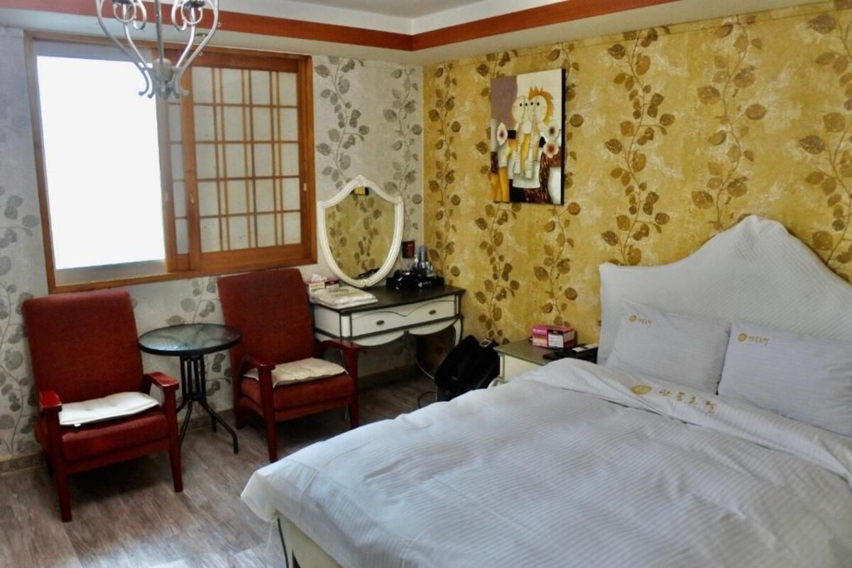 A hotel room with a bed, two chairs, a small table, a desk with a mirror, and a window with a wooden frame.