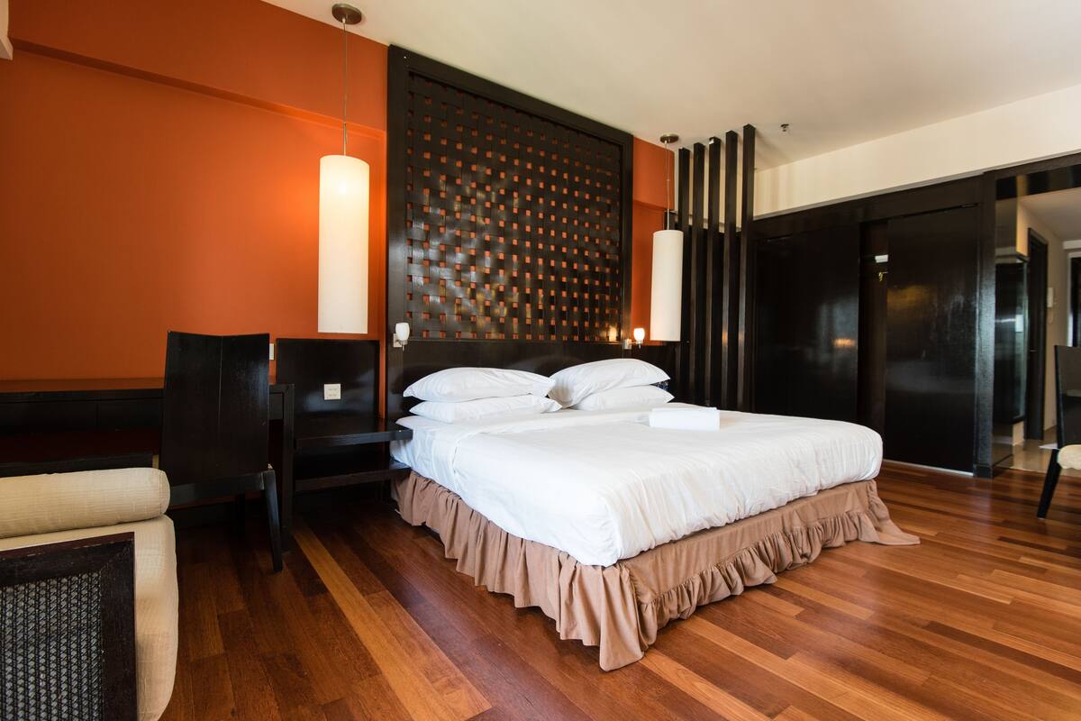 A modern hotel room with a large bed, wooden flooring, a decorative wall panel, and a seating area with a chair.