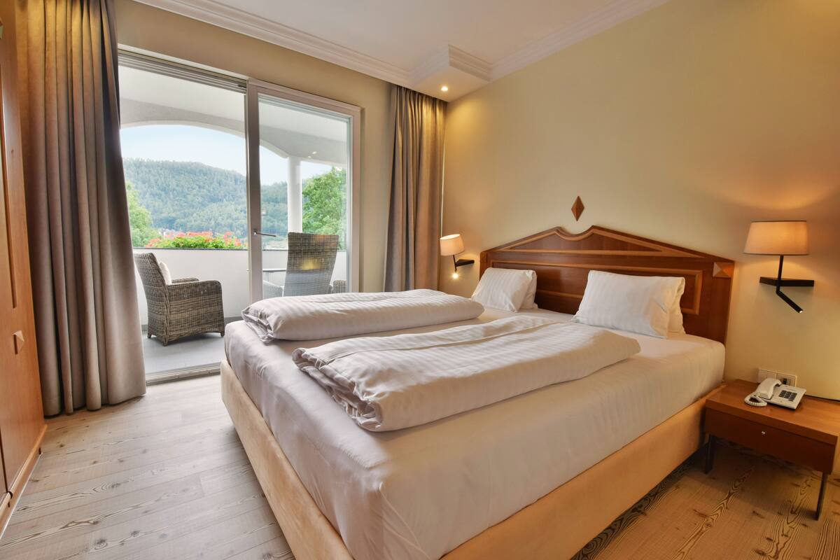 Habitación de hotel con una cama grande, una silla junto a la ventana y un balcón con vistas a árboles y colinas.