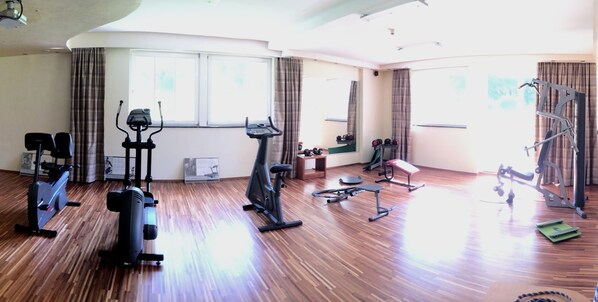 Amplia habitación con piso de madera, equipada con varias máquinas de ejercicio, incluyendo una bicicleta estática, elíptica y banco de pesas.