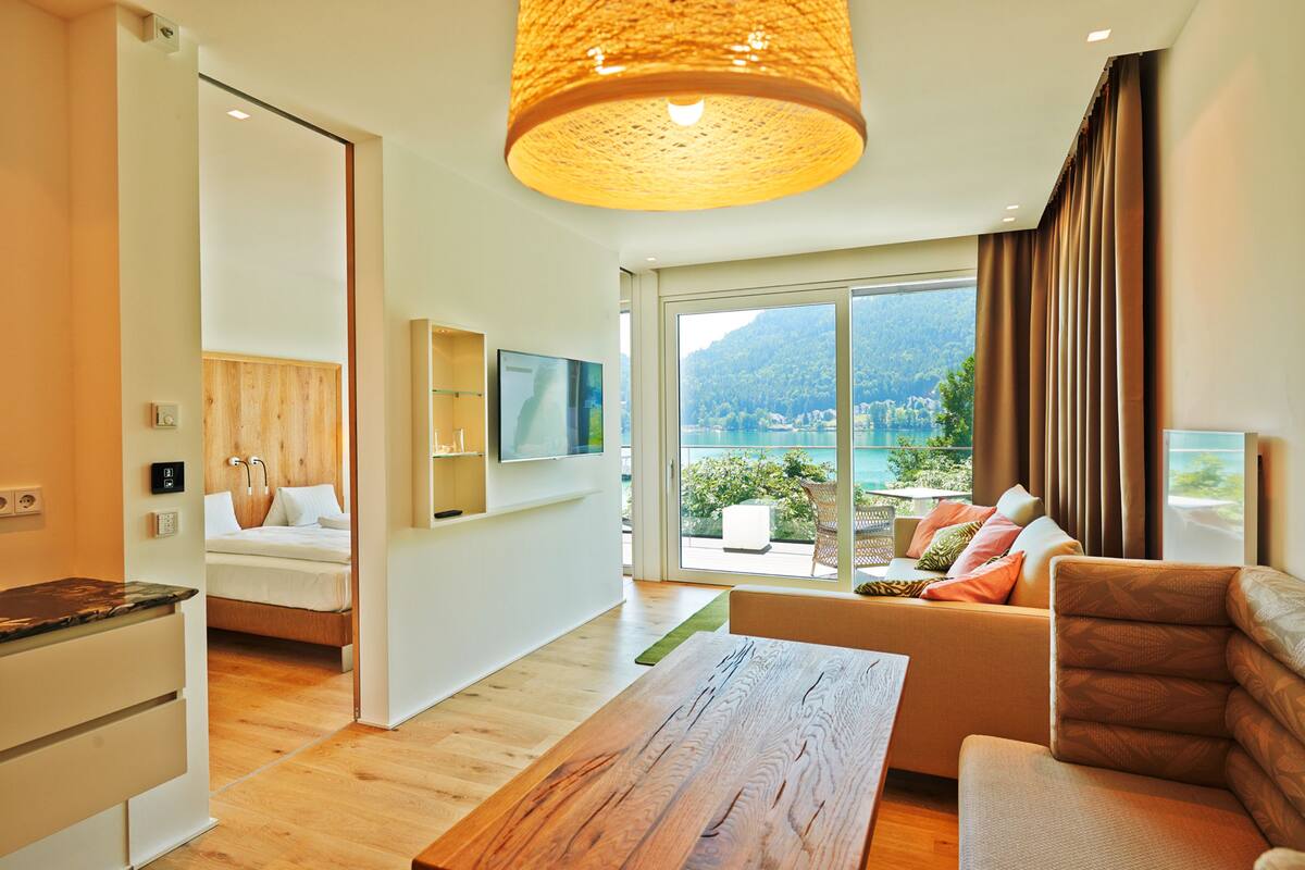 Una sala de estar moderna con piso de madera, un sofá con cojines, una mesa de centro y un amplio ventanal con vistas a un paisaje montañoso.