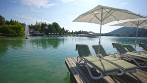 Eine Seeterrasse mit Liegestühlen und Sonnenschirmen, ein Steg mit einem Boot und ein bewaldeter Hang im Hintergrund.