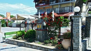 A stone pathway leading to a small church-like structure with a cross, surrounded by flower beds and a large potted plant. - Studios Pieria (Dio-Olympos)