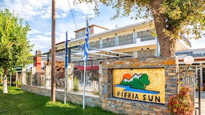 A sign for Pieria Sun with a mountain and sun graphic, surrounded by stone walls and a metal fence. - Studios Pieria (Dio-Olympos)