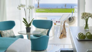 Habitación de hotel moderna con vista al océano, un balcón con mesa y sillas, y una zona de comedor con ventilador y un jarrón de flores.