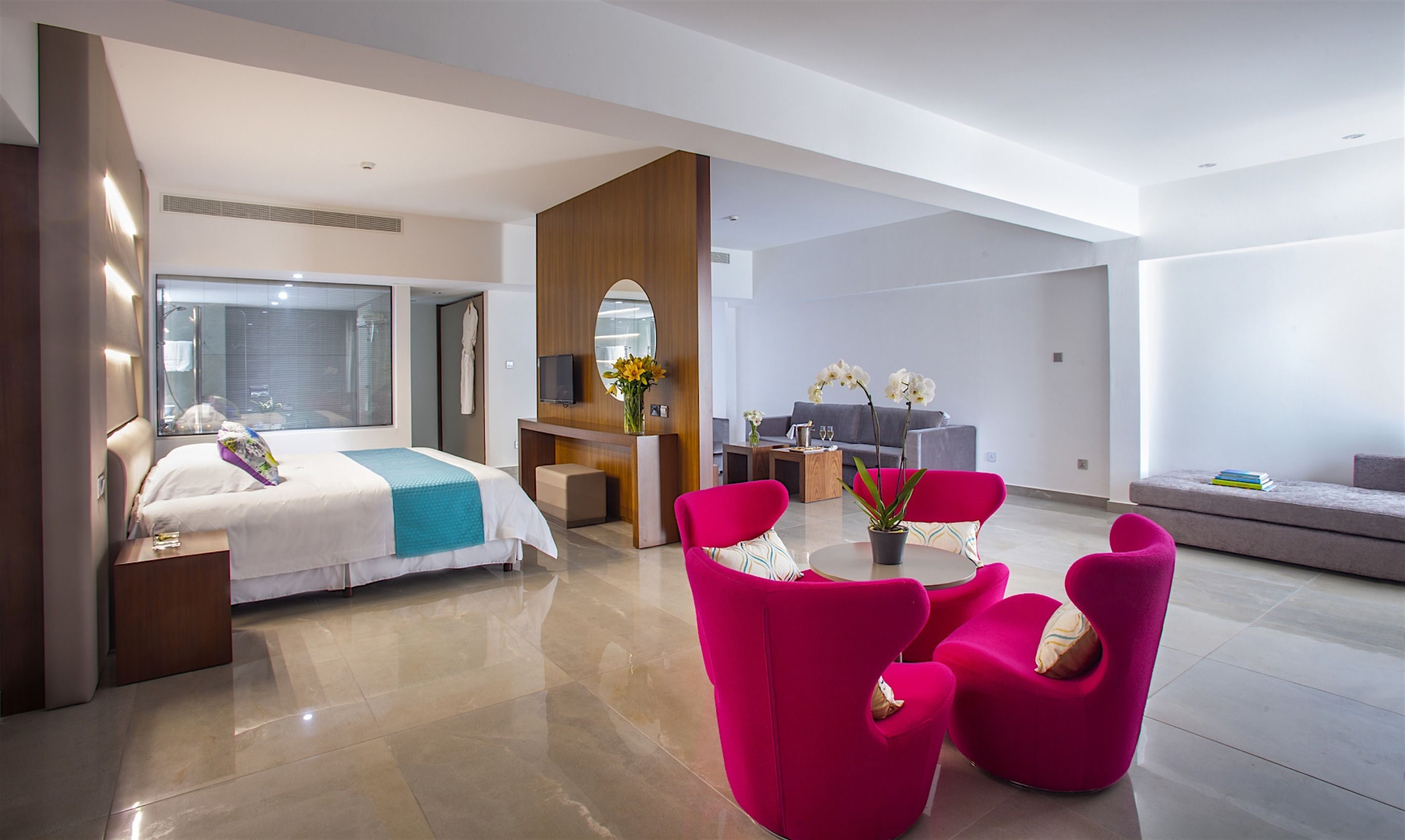 A modern hotel room with a bed, a desk, a sofa, and bright pink chairs.