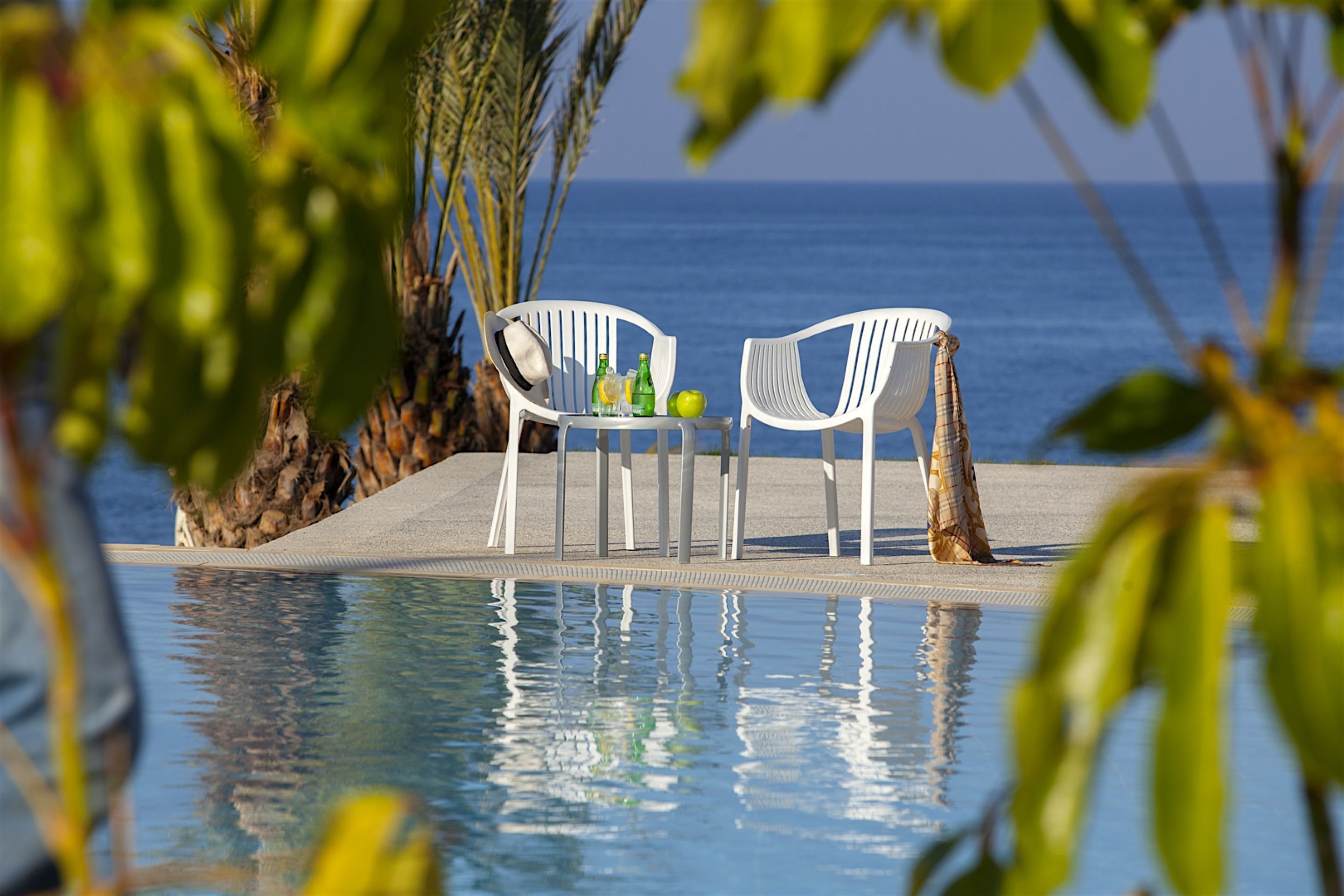 A poolside area with two white chairs and a table, a palm tree, and a view of the ocean.
