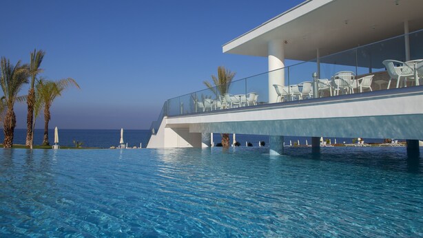 Un hotel moderno con zona de piscina, sillones blancos y sombrillas, con vistas al mar azul y cristalino.