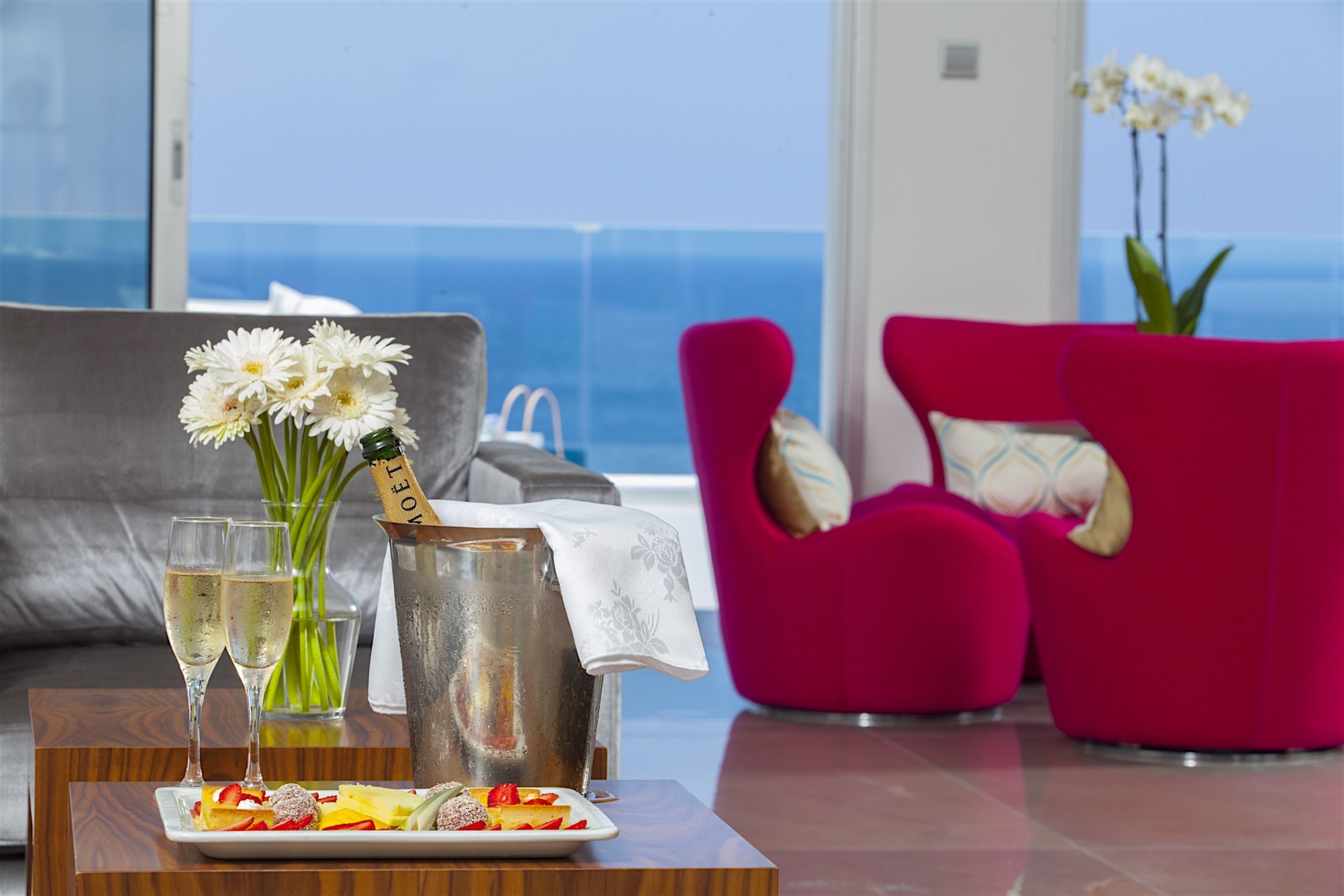 Een moderne woonkamer met een grijze bank, een roze fauteuil, een dienblad met fruit en champagne, en uitzicht op de oceaan.