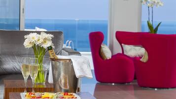Un salon moderne avec un canapé gris, un fauteuil rose, un plateau avec des fruits et du champagne, et une vue sur l’océan.