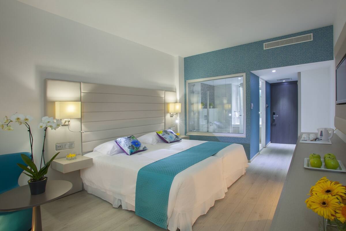 Moderni hotellihuone, jossa on suuri sänky, yöpöydällä lamput, pieni pöytä, jossa on kasvi, näkymä kylpyhuoneeseen ja pieni keittotila.