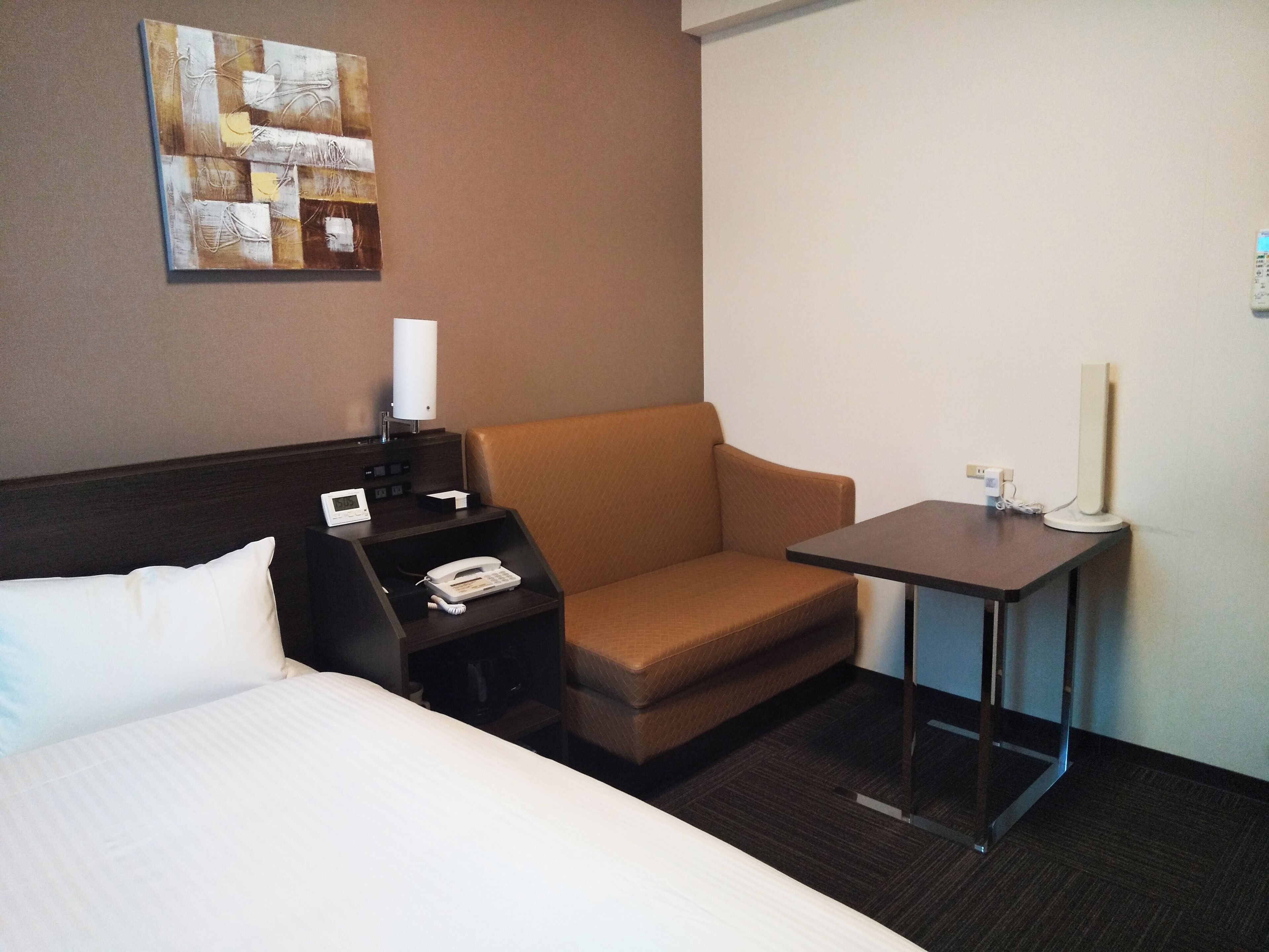Una habitación de hotel con cama, escritorio con teléfono, lámpara y una mesita.
