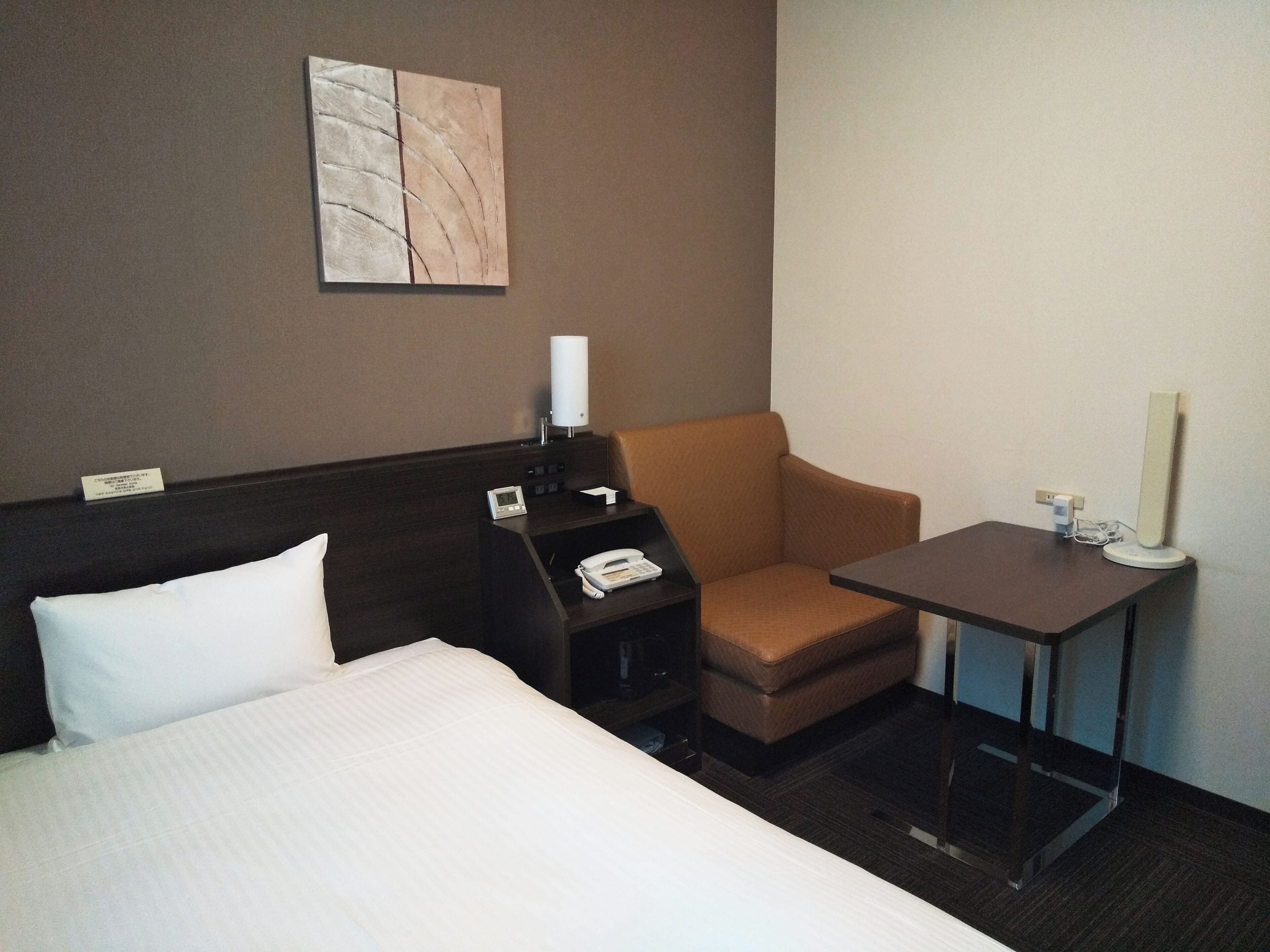 A hotel room with a bed, a desk with a telephone, a chair, a small table with a lamp, and a wall-mounted artwork.