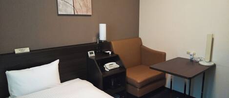 A hotel room with a bed, a desk with a telephone, a chair, a small table with a lamp, and a wall-mounted artwork.
