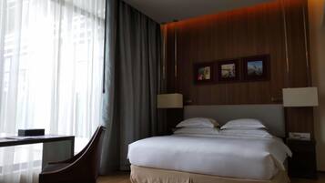 A hotel room with a large bed, a desk with a chair, and a window with sheer curtains.